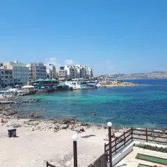 Słoneczny dzień na Bugibba beach. Ludzie odpoczywają na piasku i w turkusowej wodzie, w tle budynki i morze.