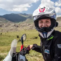 Uśmiechnięta motocyklistka w kasku, gotowa do podróży. W tle górzysty krajobraz, idealny do przygód z **carnet de passage**.