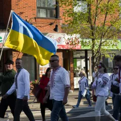 Ludzie w tradycyjnych strojach niosą flagę Ukrainy. W tle sklepy, symbolizujące życie codzienne, które wciąż trwa, mimo trudności zagrażających liczba ludności Ukrainy.