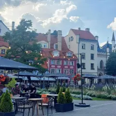 Tętniący życiem plac w Rydze, z kolorowymi kamienicami i kawiarnianymi ogródkami. W tle widać fragmenty zabytkowej architektury, przypominającej o historii państwa z Kurlandią.