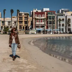 Kobieta spaceruje po piaszczystej plaży w pięknym, słonecznym dniu. W tle kolorowe budynki i palmy tworzą urokliwy krajobraz.