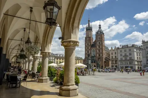 Urokliwy **Kraków Stary Miasto** z Sukiennicami i Kościołem Mariackim w tle. Widać kawiarniane stoliki pod arkadami.
