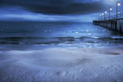 Nocna, świecąca plaża z molo i latarniami w oddali. Piasek lśni w księżycowym blasku.
