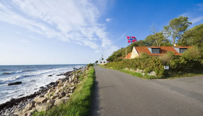 Droga nad morzem, domy z norweską flagą. Idealne miejsce na wycieczkę rowerem na Bornholm, warto sprawdzić koszty.