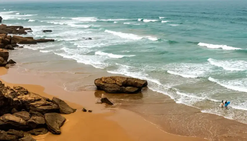 Maroko plaże: skaliste wybrzeże, piaszczysta plaża i turkusowe fale Atlantyku. Dwóch surferów wchodzi do wody z deskami.