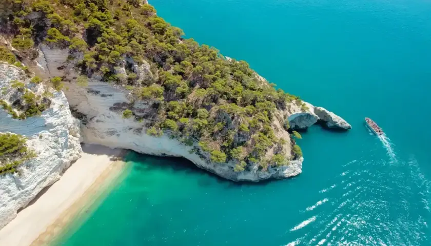 Widok z lotu ptaka na skaliste wybrzeże z piaszczystą plażą i turkusową wodą. Statek wycieczkowy płynie w stronę łuku skalnego. Idealne miejsce, gdzie do Włoch!