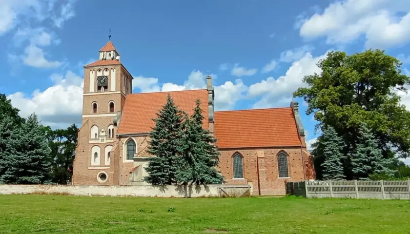 Nieszawa atrakcje: ceglany kościół z wieżą i zegarem, otoczony zielenią, pod błękitnym niebem z chmurami.