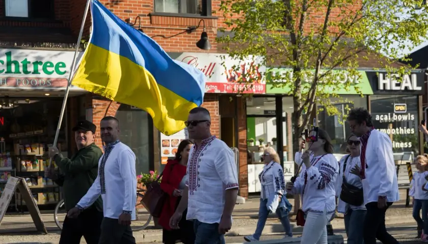 Ludzie w tradycyjnych strojach niosą flagę Ukrainy. W tle sklepy, symbolizujące życie codzienne, które wciąż trwa, mimo trudności zagrażających liczba ludności Ukrainy.