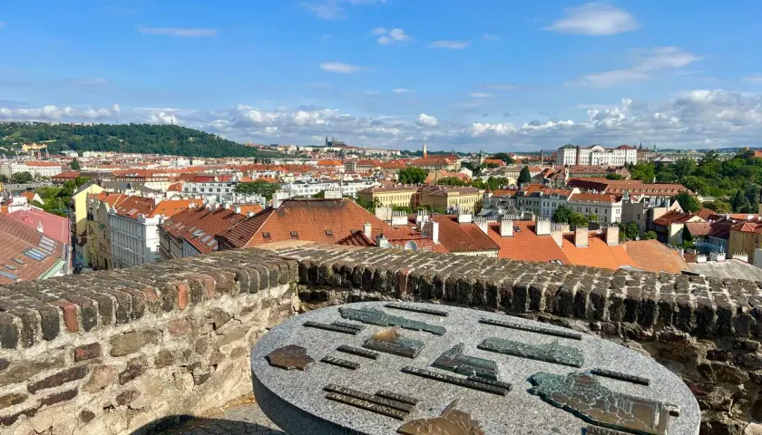 Widok na Pragę z Wyszehradu. Na pierwszym planie kamienny stół z mapami zabytków. W tle czerwone dachy i zielone wzgórza.