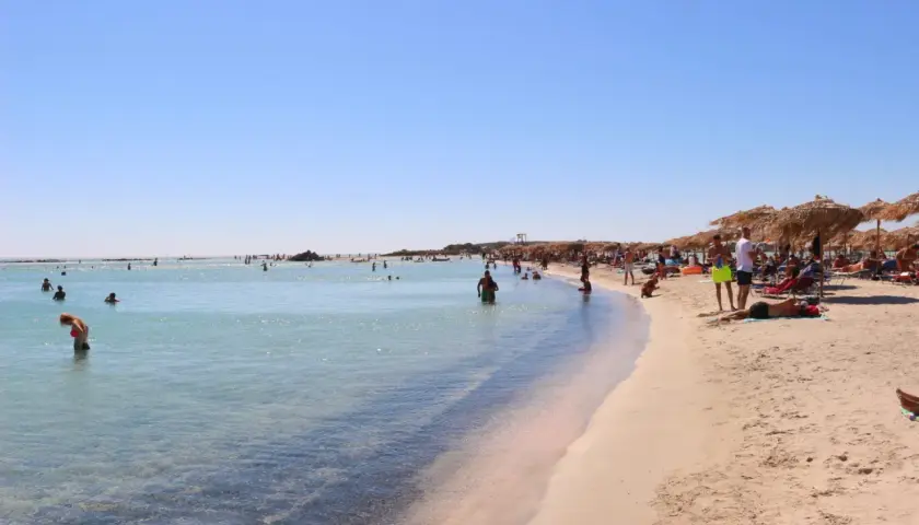 Słoneczna plaża Elafonisi z turkusową wodą, piaskiem i ludźmi odpoczywającymi pod parasolami.