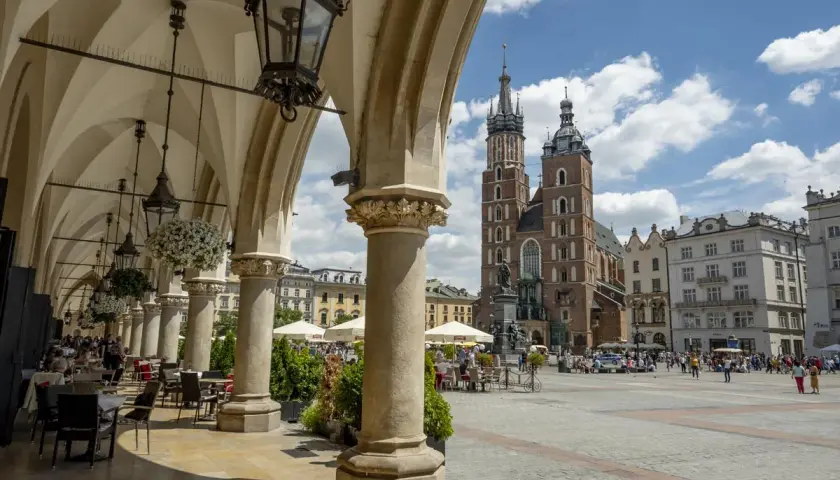 Urokliwy **Kraków Stary Miasto** z Sukiennicami i Kościołem Mariackim w tle. Widać kawiarniane stoliki pod arkadami.