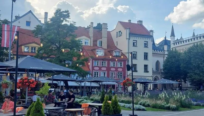 Tętniący życiem plac w Rydze, z kolorowymi kamienicami i kawiarnianymi ogródkami. W tle widać fragmenty zabytkowej architektury, przypominającej o historii państwa z Kurlandią.