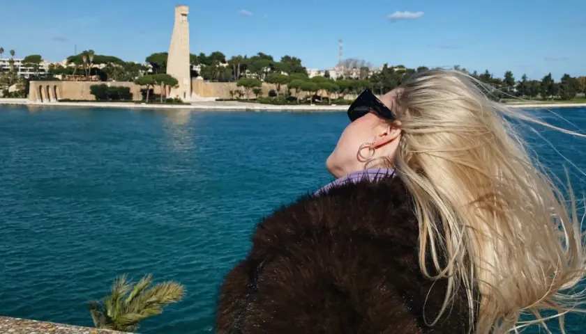 Widok na pomnik i morze w Brindisi. Wiatr rozwiewa blond włosy kobiety w futrze.