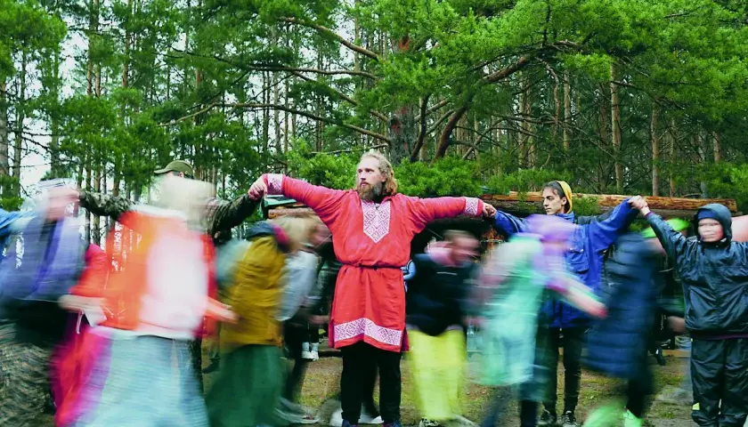 Ludzie w lesie tańczą w kręgu. W centrum mężczyzna w czerwonej koszuli. To obraz życia, a nie statystyka rosja liczba ludności.