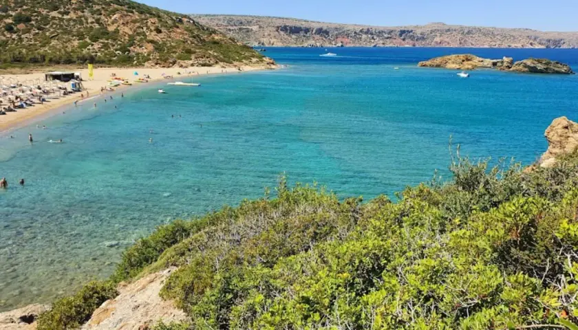 Słoneczna plaża Vai z turkusową wodą, zielonymi wzgórzami i ludźmi relaksującymi się na piasku.