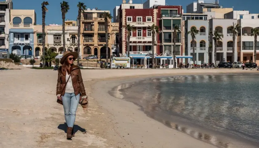Kobieta spaceruje po piaszczystej plaży w pięknym, słonecznym dniu. W tle kolorowe budynki i palmy tworzą urokliwy krajobraz.
