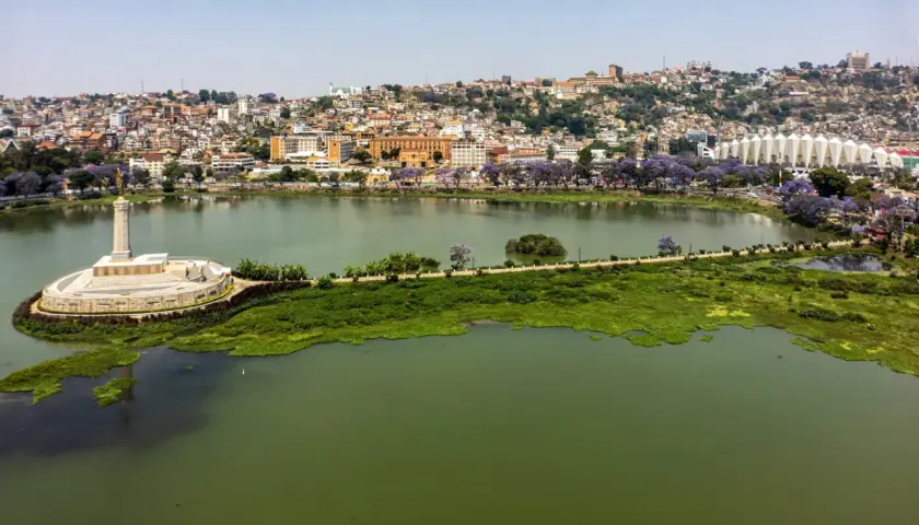 Widok na jezioro i wzgórza Antananarywy, gdzie żyje madagaskar ludność. Widać pomnik, stadion i kwitnące drzewa.