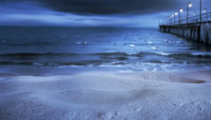 Nocna, świecąca plaża z molo i latarniami w oddali. Piasek lśni w księżycowym blasku.