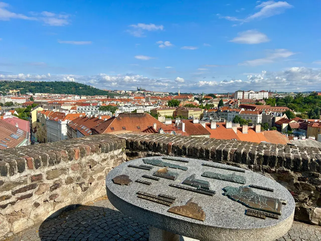 Widok na Pragę z Wyszehradu. Na pierwszym planie kamienny stół z mapami zabytków. W tle czerwone dachy i zielone wzgórza.
