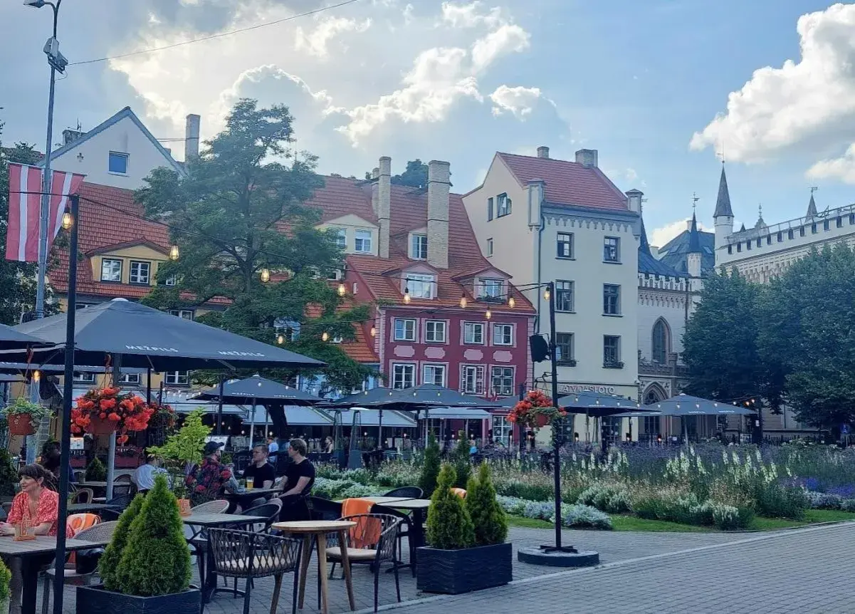 Tętniący życiem plac w Rydze, z kolorowymi kamienicami i kawiarnianymi ogródkami. W tle widać fragmenty zabytkowej architektury, przypominającej o historii państwa z Kurlandią.